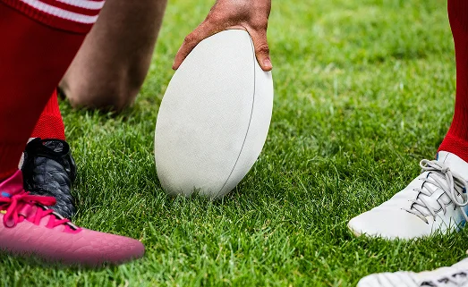 Gros plan sur des joueurs de rugby en train de placer le ballon sur la pelouse pour le coup d'envoi, on voit leurs crampons et leurs mains alors qu'ils se préparent à jouer