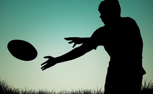 Silhouette d'un joueur de rugby en train de passer le ballon sur un terrain en gazon au coucher du soleil, illustrant le mouvement et l'esprit d'équipe propres à ce sport